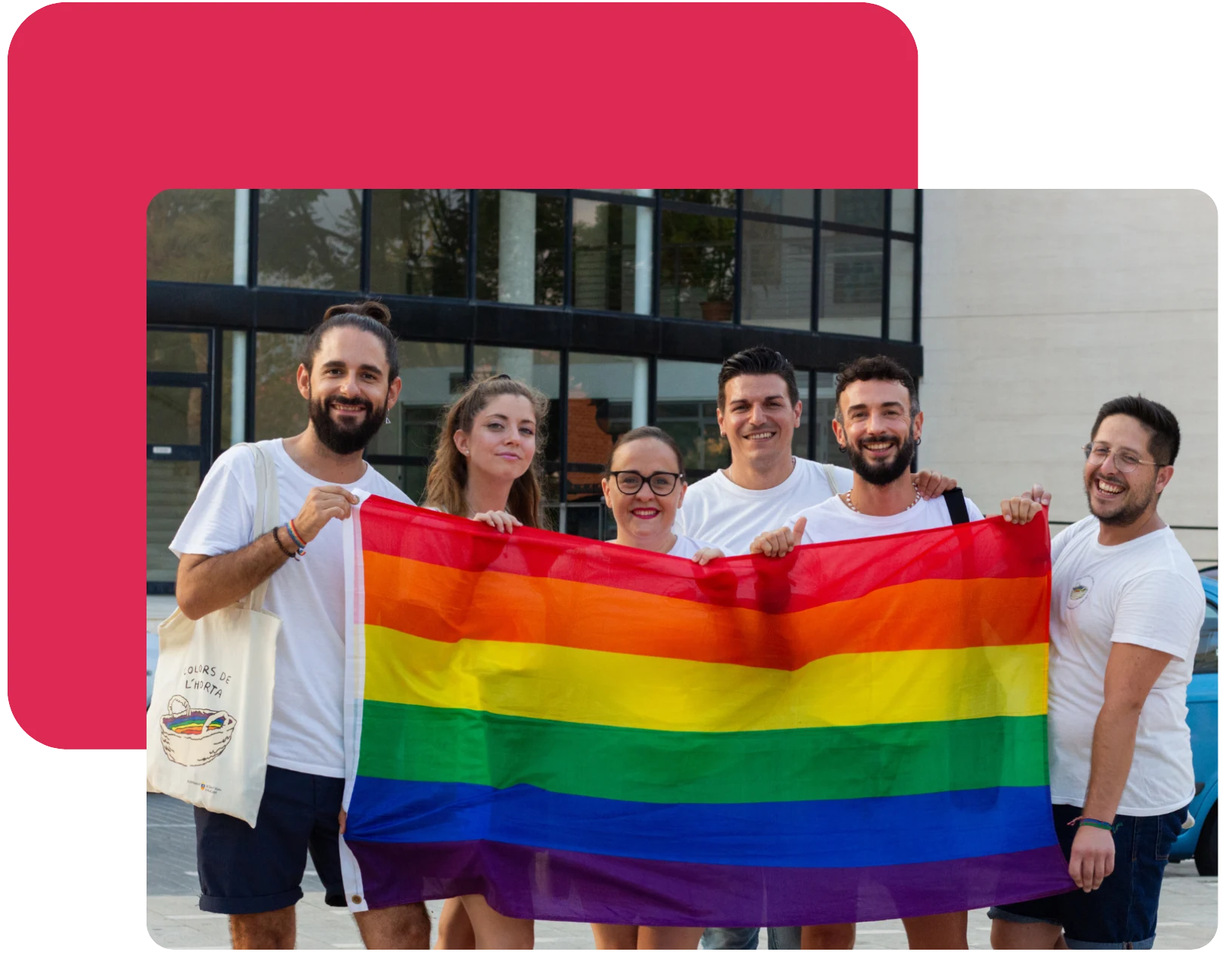 6 miembros de Colors de l'Horta sostienen sonrientes una bandera LGTBI delante de la Casa de Cultura del pueblo.