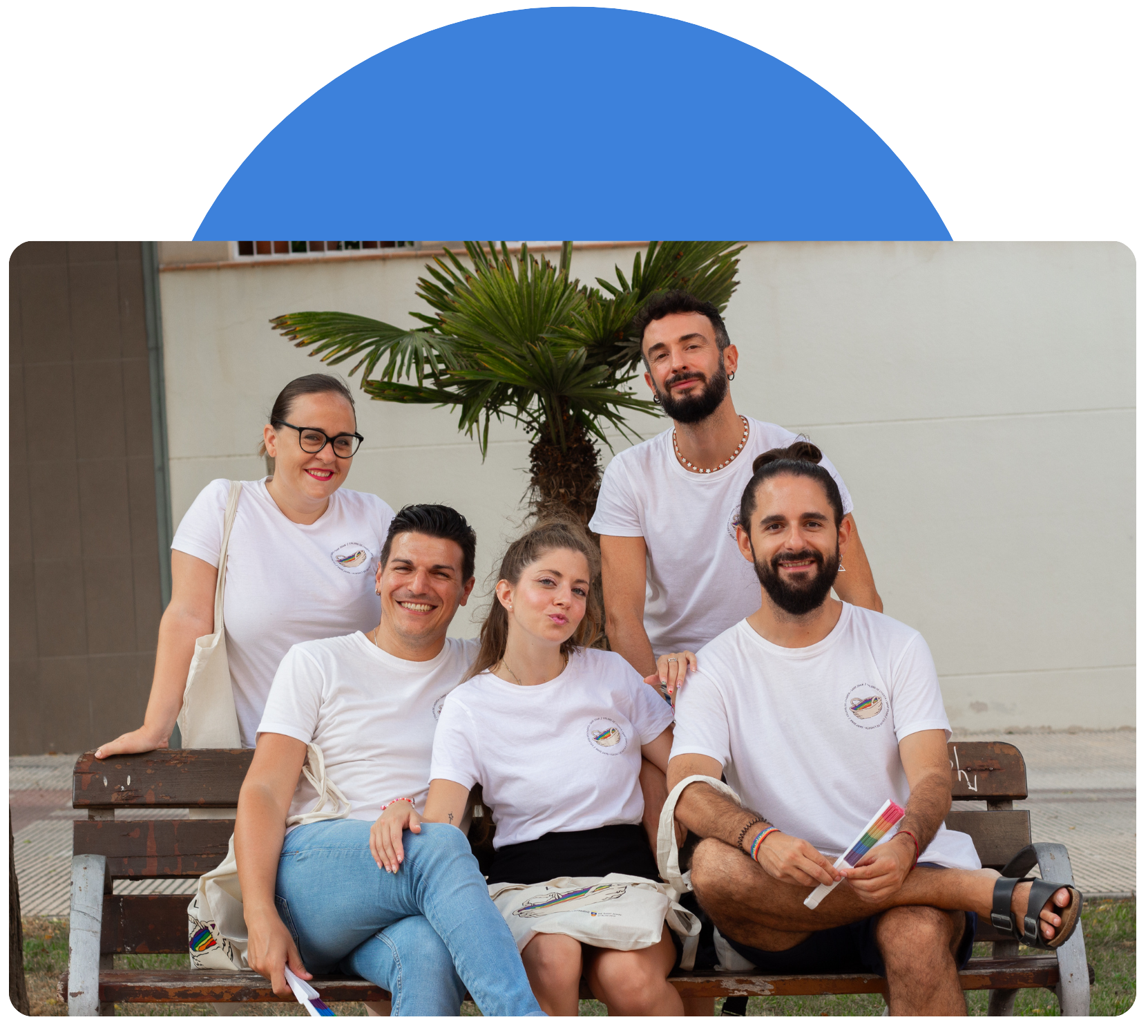 Cinco miembros de la asociación LGTBI Colors de l'Horta posan sentados en un banco del pueblo sonrientes con la camiseta de la asociación.