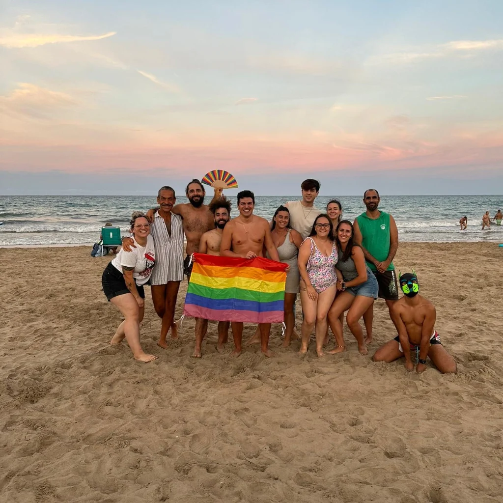 13 personas en la playa posando con una bandera LGTBI en una de las convivencias de verano de la asociación.