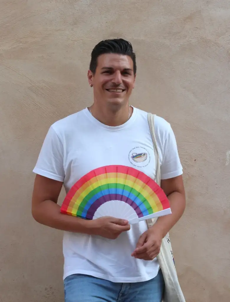 Fotografía de Sergio Agueitos. Hombre blanco de unos 30 años, delgado, pelo corto negro. Lleva la camiseta de Colors de l'Horta (blanca con el logo en pequeño) y un abanico arcoiris.